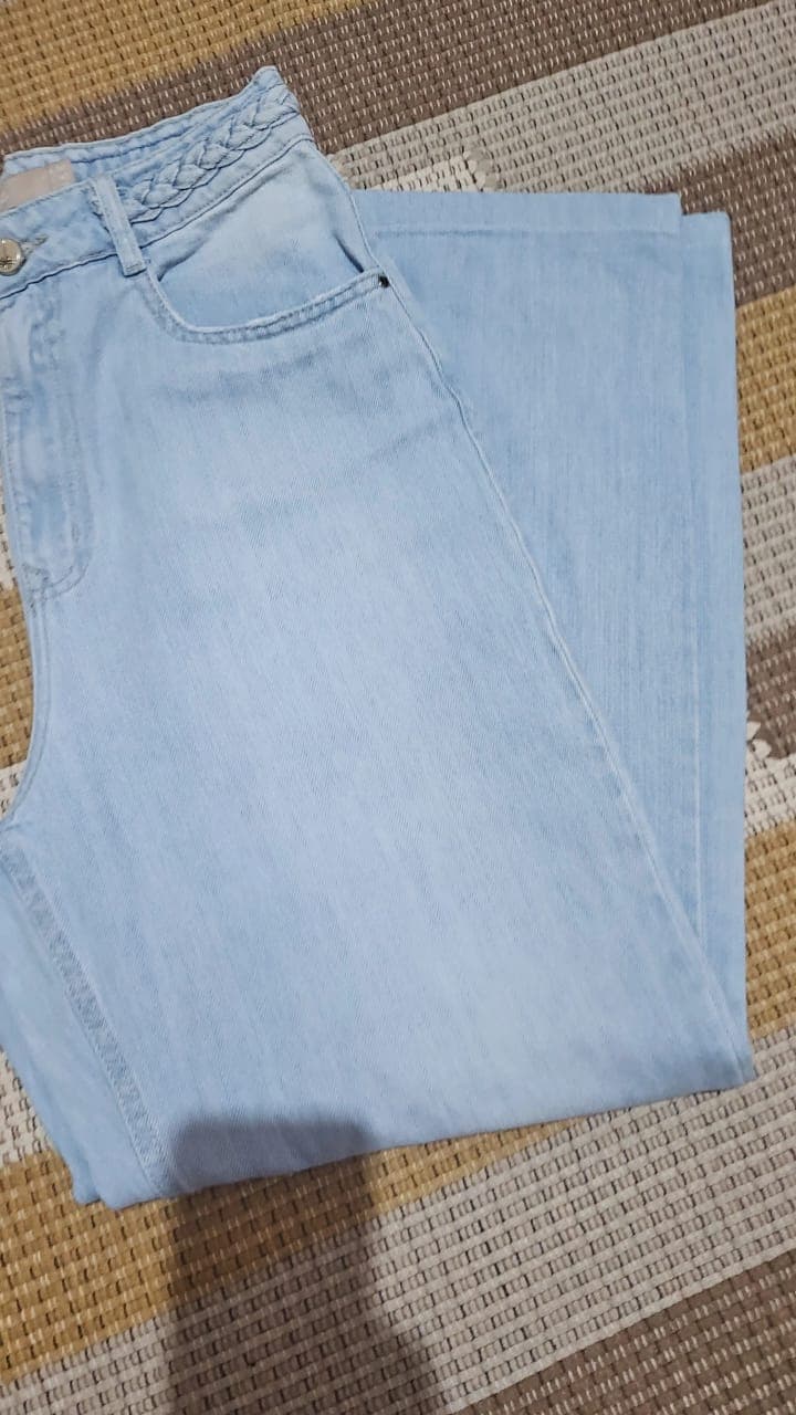 Calça jeans de lavagem azul bem clara, dobrada, mostrando o cós com detalhe trançado e a perna larga