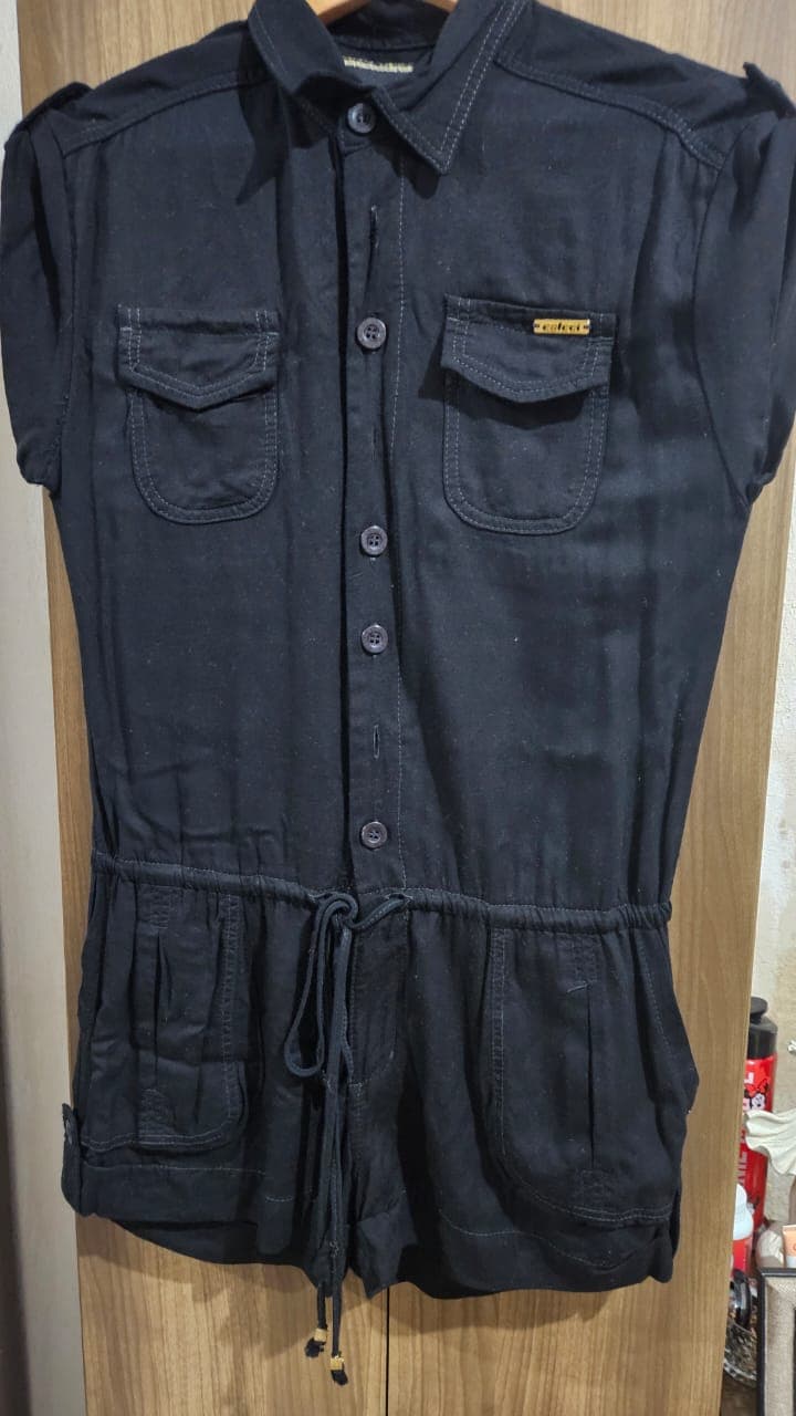 Macaquinho preto no cabide, gola de camisa, fileira de botões, dois bolsos com pala no peito, cordão amarrado na cintura e bolsos no short