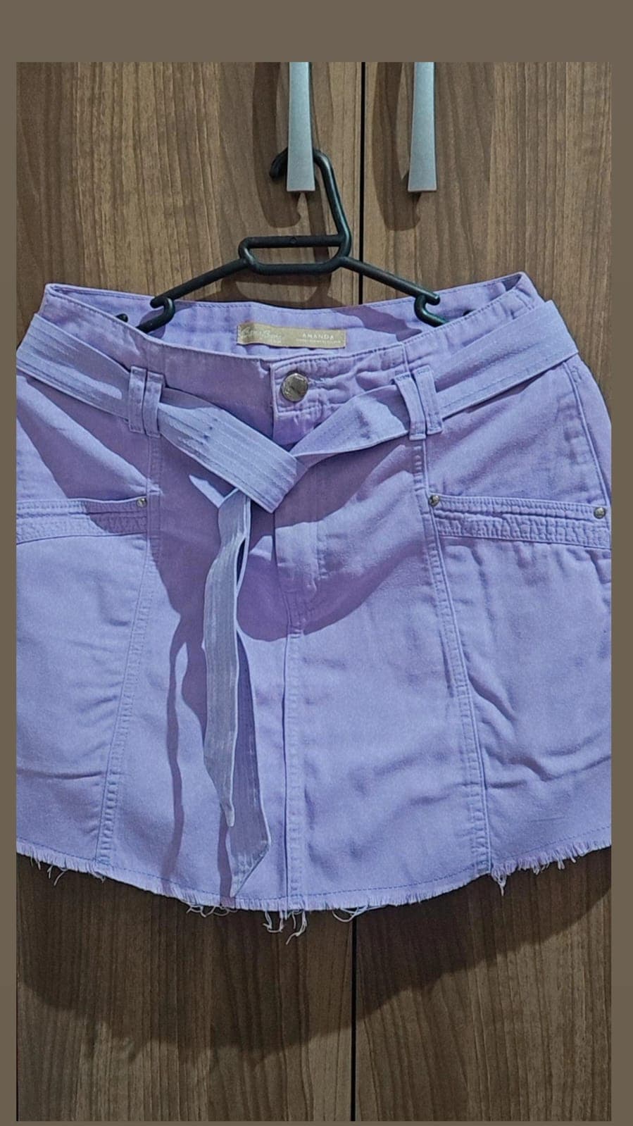 Saia jeans lilás no cabide, vista de frente, com faixa da mesma cor amarrada na cintura e barra desfiada