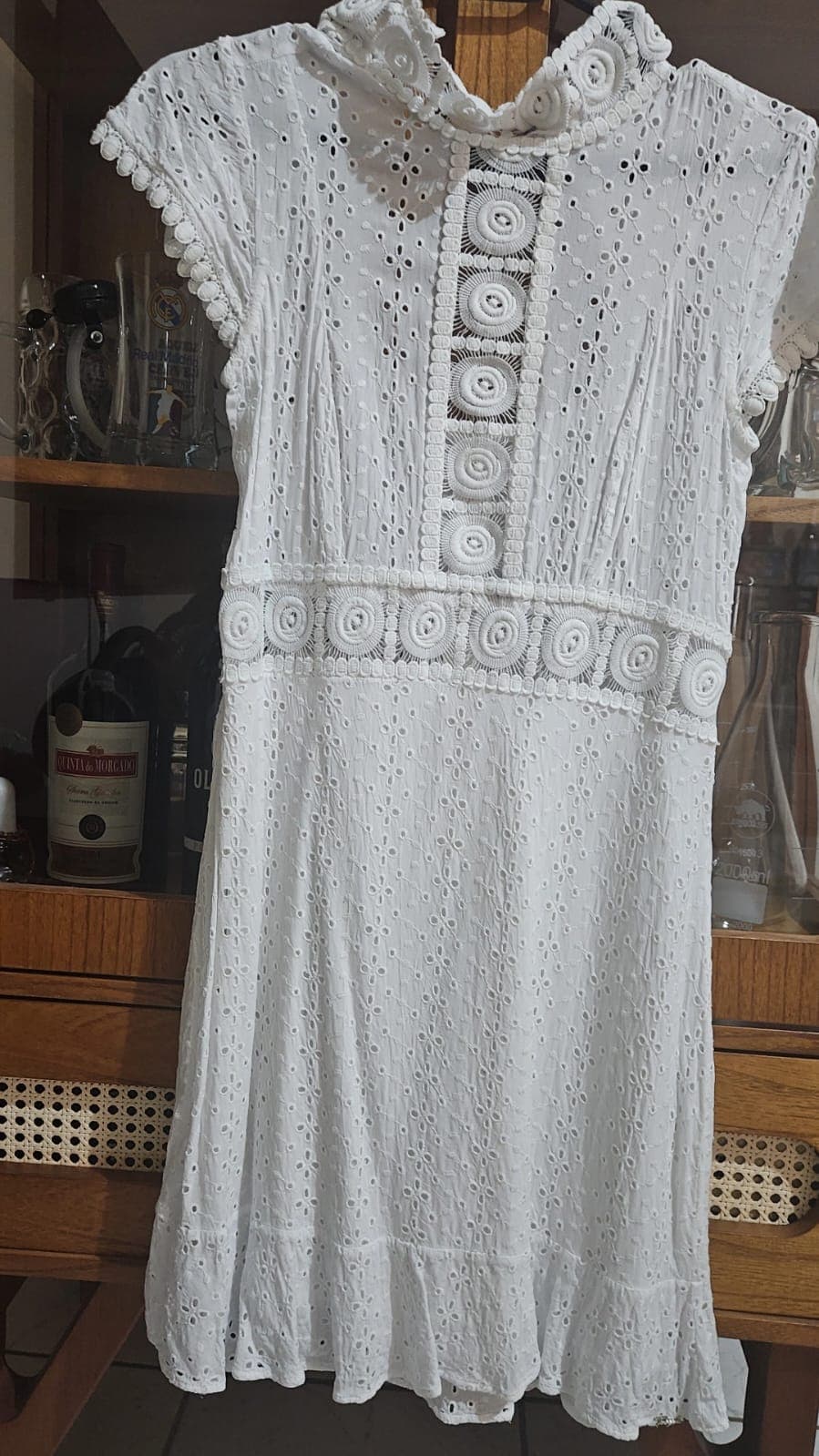 Vestido branco de laise bordada no cabide, gola alta, fileira de botões de crochê na frente, faixa de crochê na cintura e manga curta com acabamento de bolinhas
